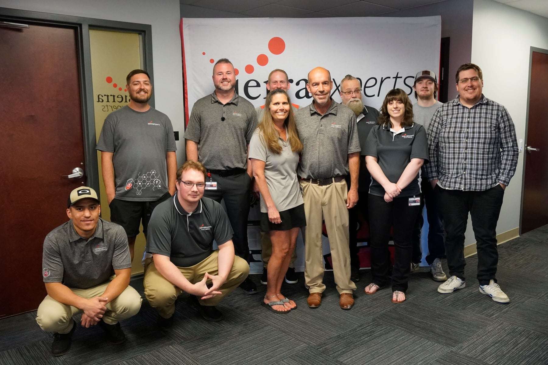 The Sierra team posed together in the office in front of a Sierra Experts banner, most in gray and black Sierra polo shirts, with a few crouching in the front row